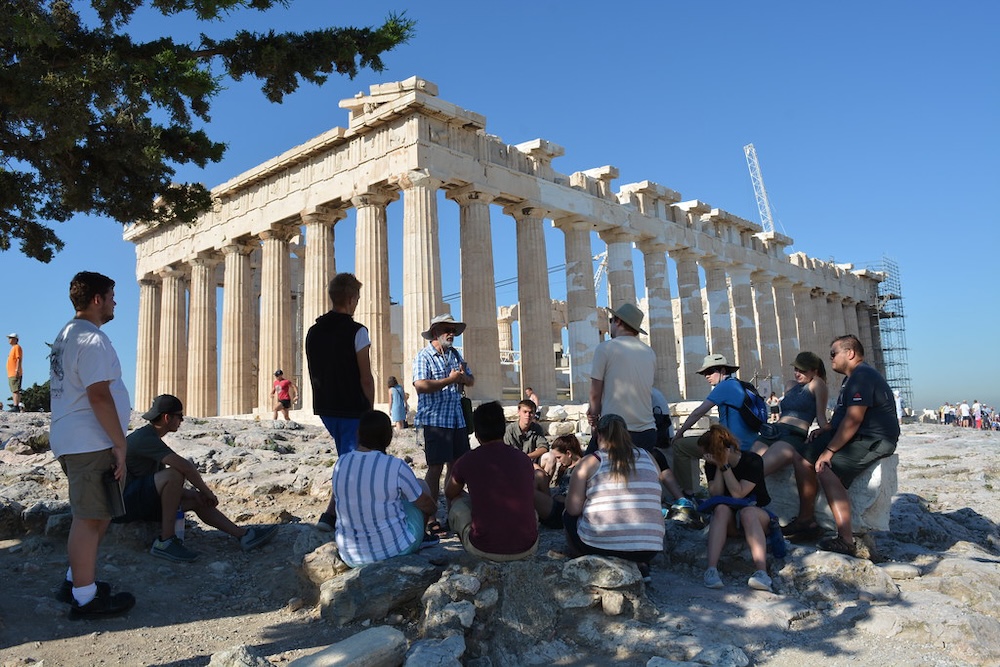 A group of people listens to a guide in front of the Parthenon temple on the Acropolis in Athens, Greece, on a clear, sunny day.