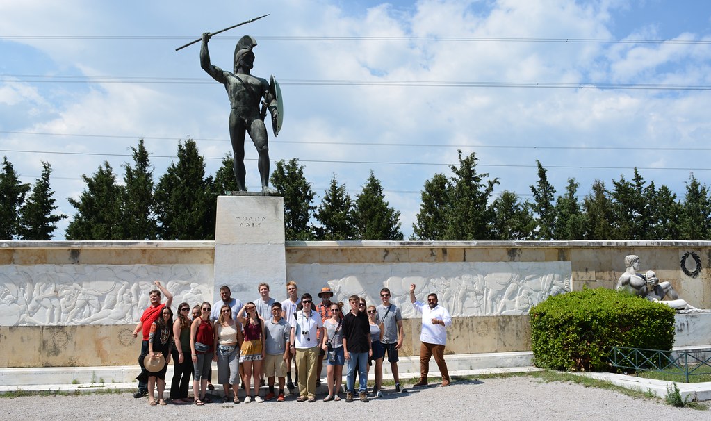 A group of people poses in front of a large bronze statue of a warrior holding a spear and shield, with a marble relief and trees in the background.