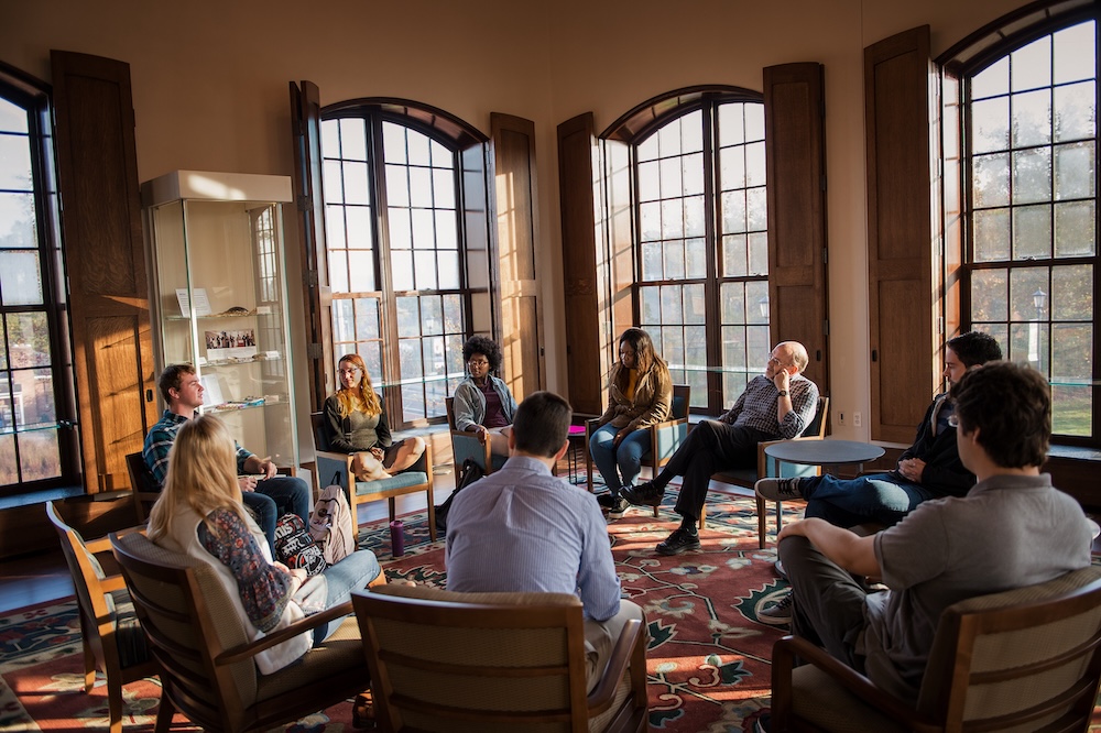 A group of people sit in a circle in a sunlit room with large windows, engaged in a discussion or meeting.