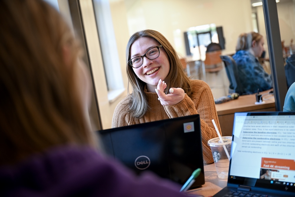 A smiling woman wearing glasses gestures while sitting at a table with a laptop and an iced drink, talking to another person.