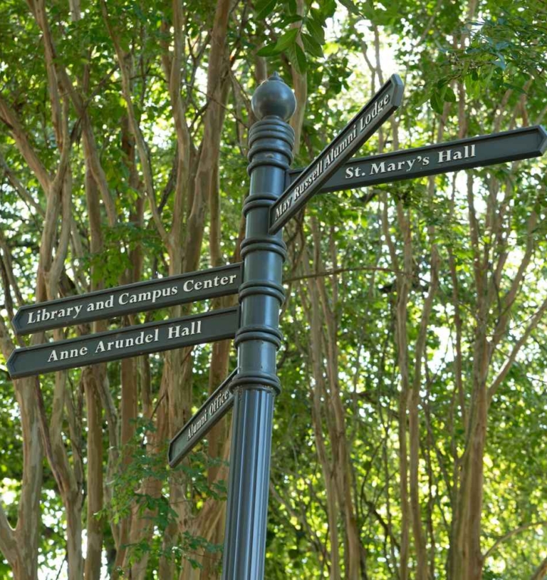 A black signpost with directional signs for Library and Campus Center, Anne Arundel Hall, and St. Mary's Hall, surrounded by green trees.