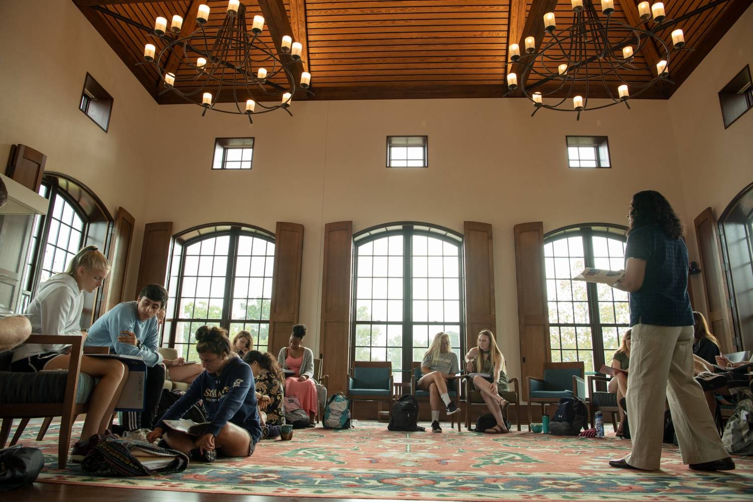 Students sit and work in a spacious room with large windows and high ceilings as a teacher stands at the front, addressing the group.