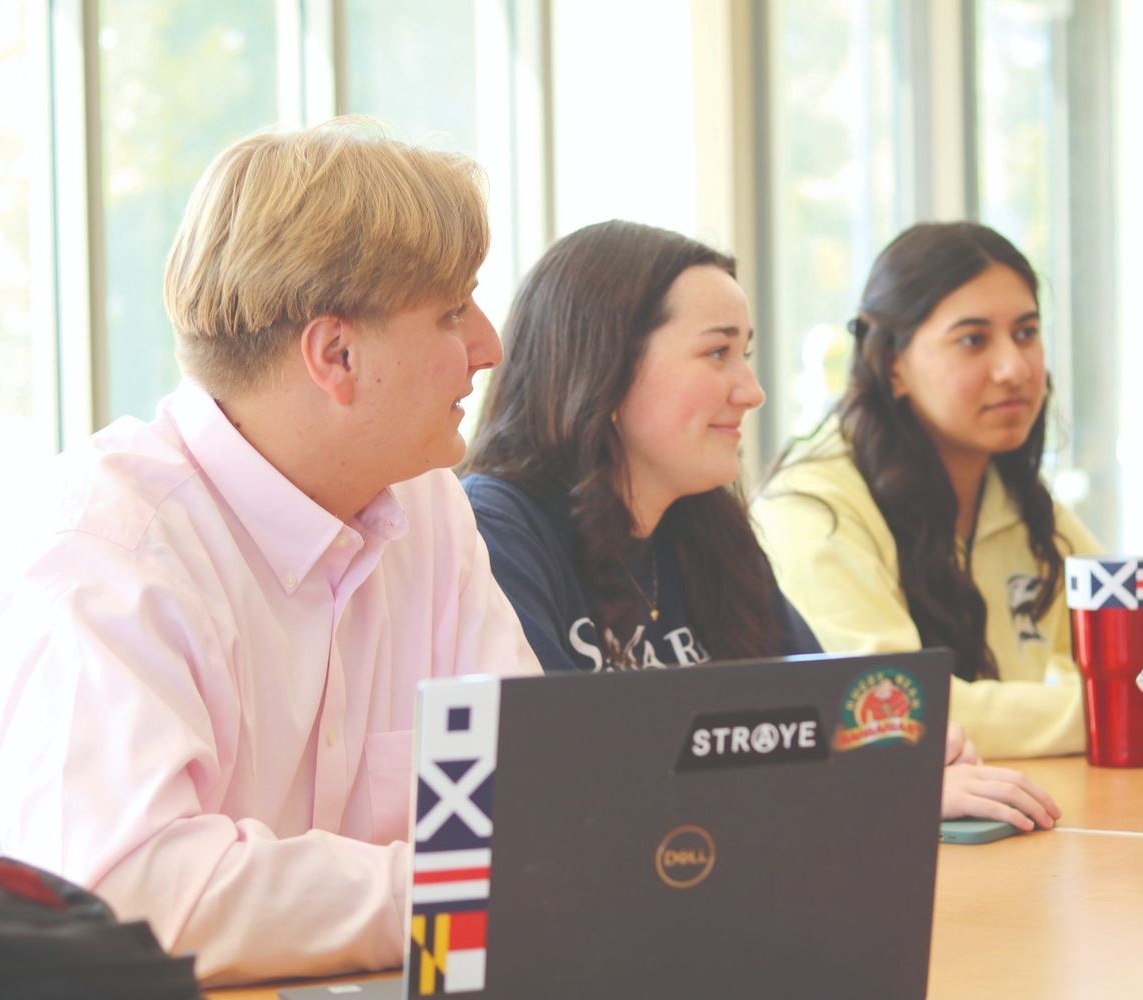 Three people sit at a table with laptops and drinks, engaged in conversation near large windows in a bright, indoor setting.