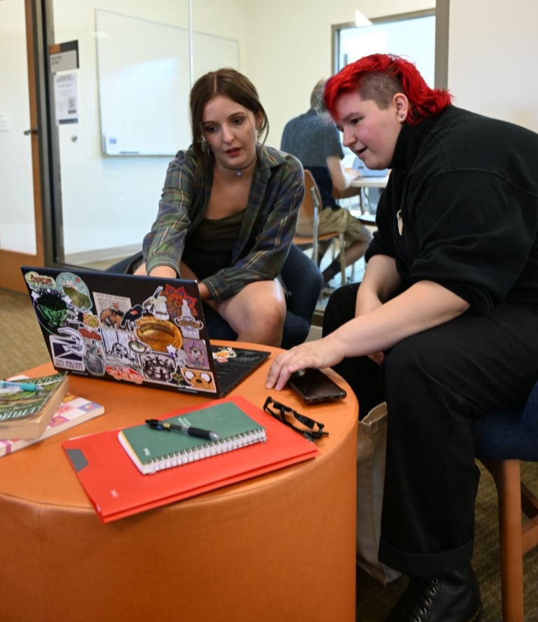 Two people sit together looking at a laptop on an orange ottoman, surrounded by notebooks and papers, in a study or office setting.