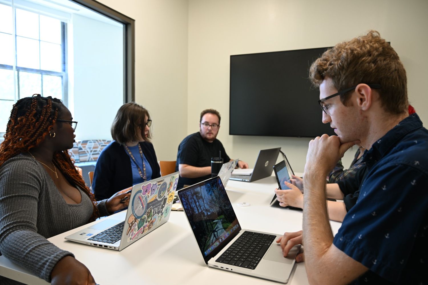 Five people sit around a table with laptops in a meeting room, engaged in discussion. A large monitor is mounted on the wall behind them.