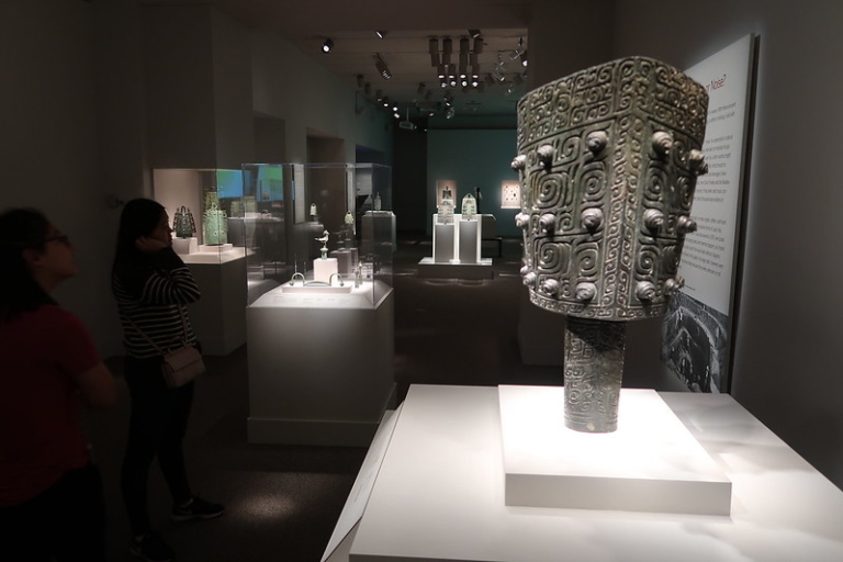 皇冠体育平台ors observe ancient bronze artifacts displayed in glass cases and on pedestals in a dimly lit museum gallery.