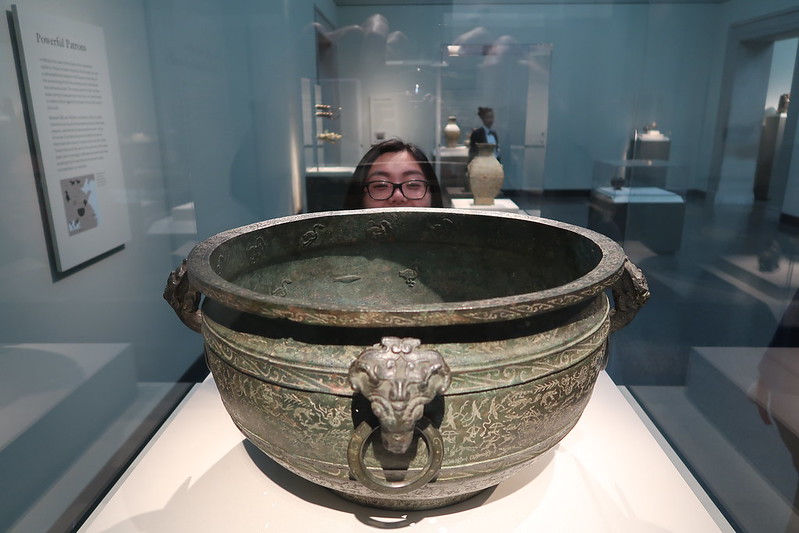 A large, ornate bronze vessel with a lion head handle is displayed in a museum; a person is partially visible behind it, peeking over the rim.