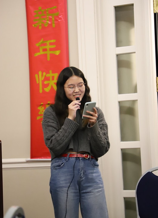 A young woman speaks into a microphone while reading from her phone, standing next to a red banner with Chinese characters.