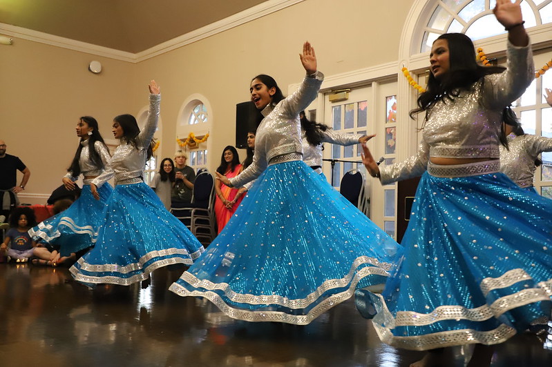A group of women wearing silver tops and blue skirts perform a synchronized dance indoors, with an audience watching in the background.