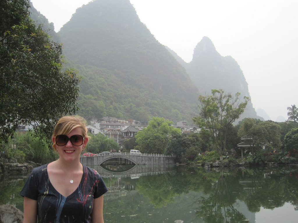A woman with sunglasses stands by a pond with a stone bridge, village buildings, and green karst mountains in the background.