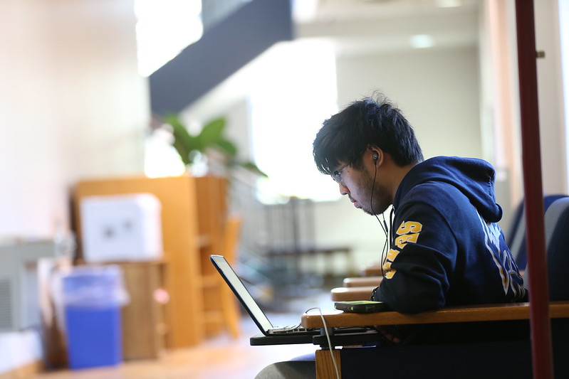 A person wearing a hoodie sits in a chair, using a laptop and listening to earphones in a well-lit indoor space.