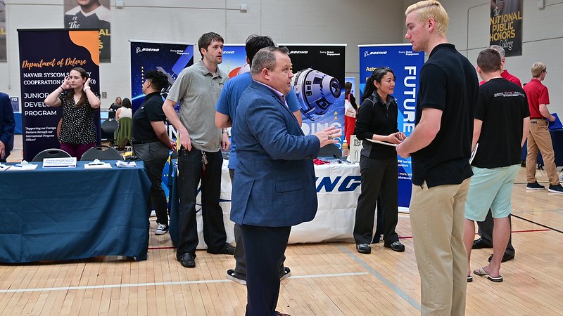 People interact at a career fair in a gymnasium, with company booths and informational banners in the background.