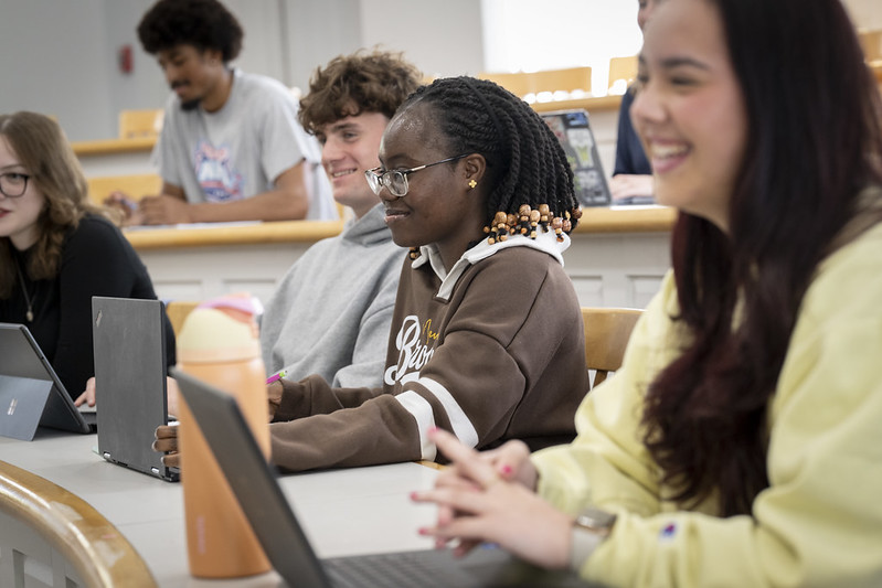Students sit at desks in a classroom, using laptops and smiling during what appears to be a group activity or lecture.