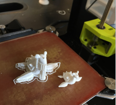 Two partially finished white 3 d printed models sit on a printer bed, with one appearing incomplete or detached; parts of the 3 d printer are visible in the background.
