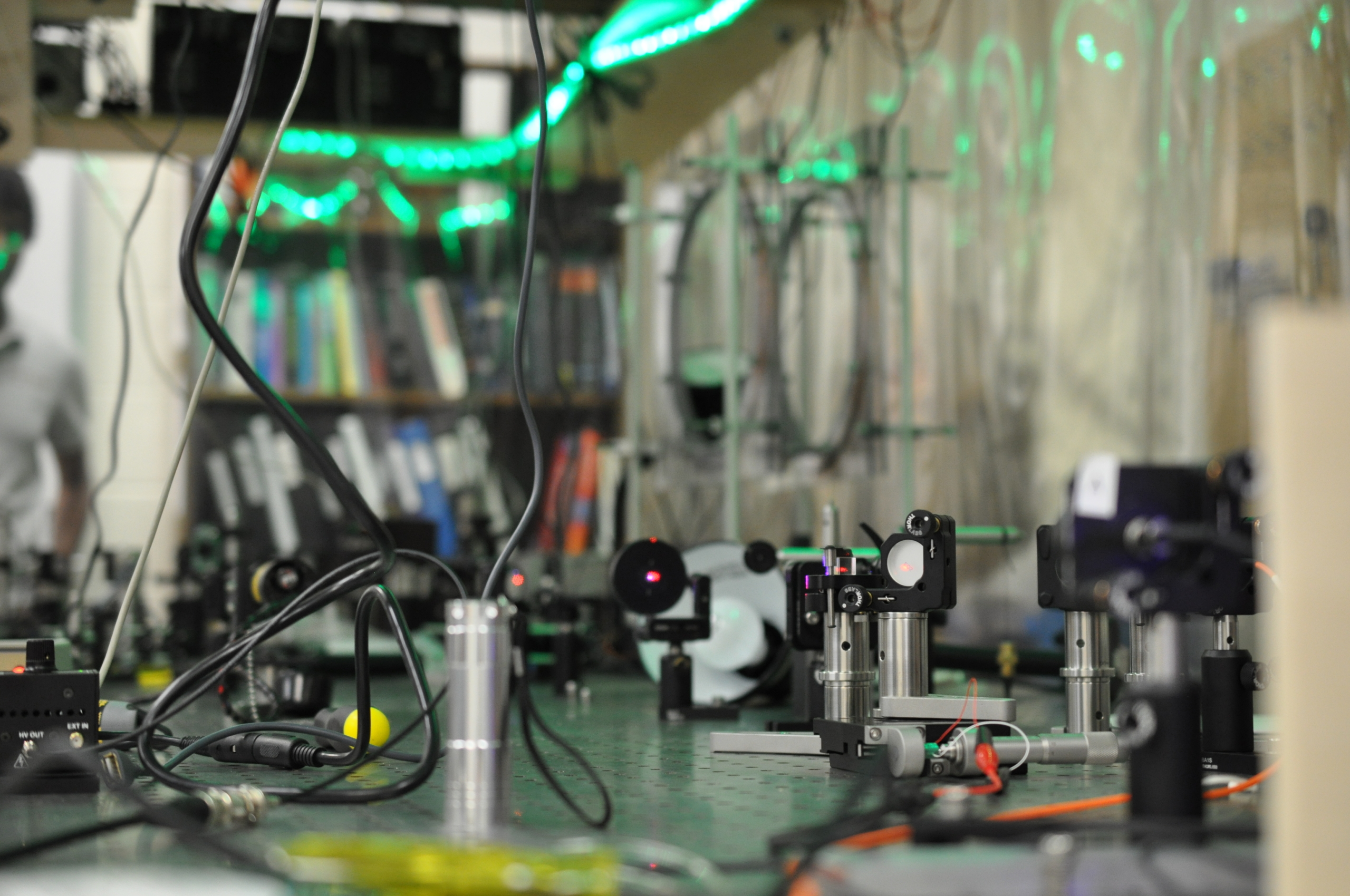 A laboratory optical table with various scientific instruments, lenses, and wires, with green LED lights and shelves of books in the background.