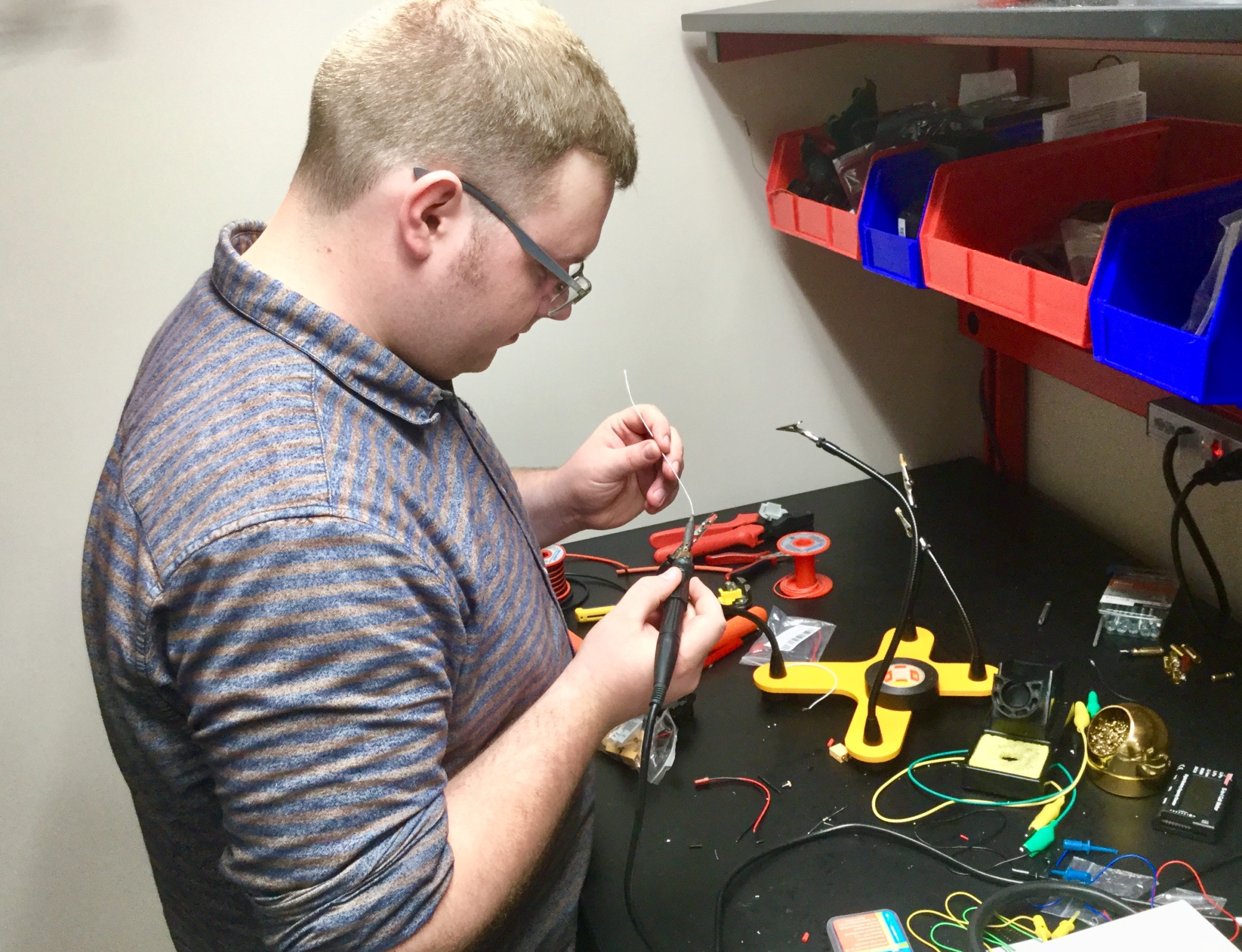 A person soldering wires at a workbench with electronic components, tools, and parts organized in bins and trays.