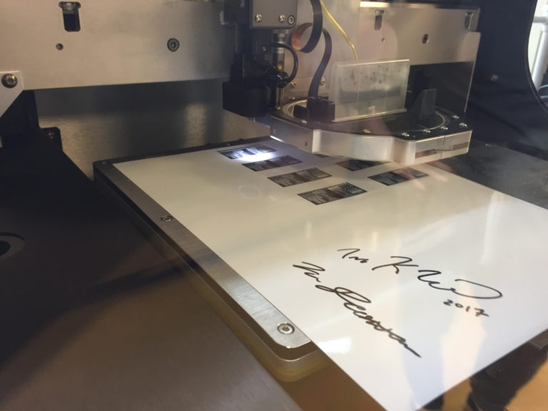 A machine is printing black rectangular shapes and two signatures dated 2017 on a white sheet.