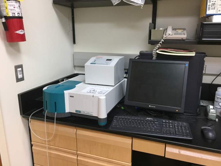 A laboratory workstation with a benchtop analyzer, computer monitor, keyboard, mouse, and office phone on a black counter.
