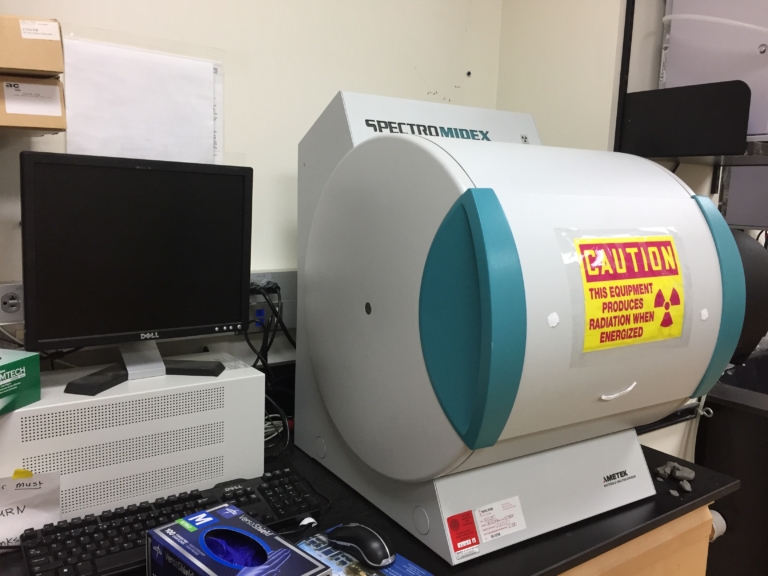 A SpectroMidex machine with a warning label is set up next to a Dell computer monitor and tower on a laboratory desk.