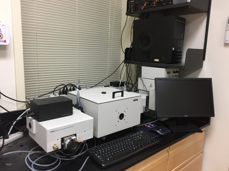 A laboratory setup featuring a spectrofluorometer, a computer monitor, keyboard, and various electronic equipment on a black countertop near window blinds.