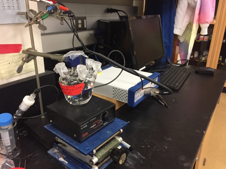 Laboratory setup with glassware on a magnetic stirrer, computer equipment, and lab supplies on a black countertop.
