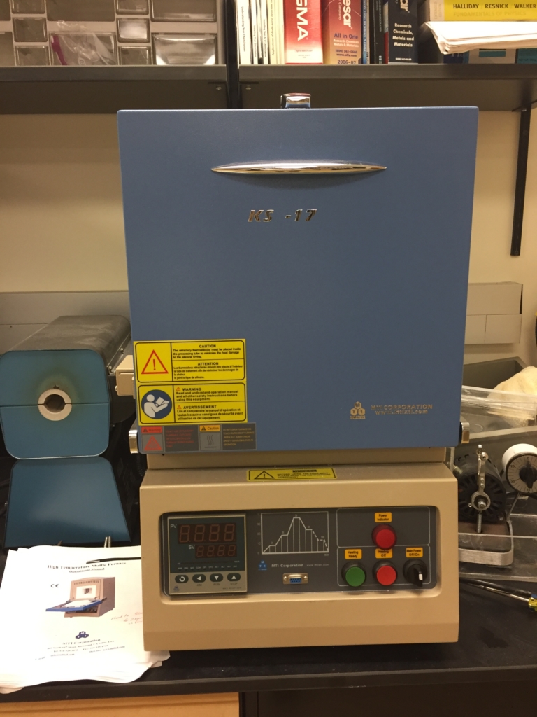 A laboratory muffle furnace with a blue chamber and digital control panel is placed on a black countertop, surrounded by lab equipment and manuals.