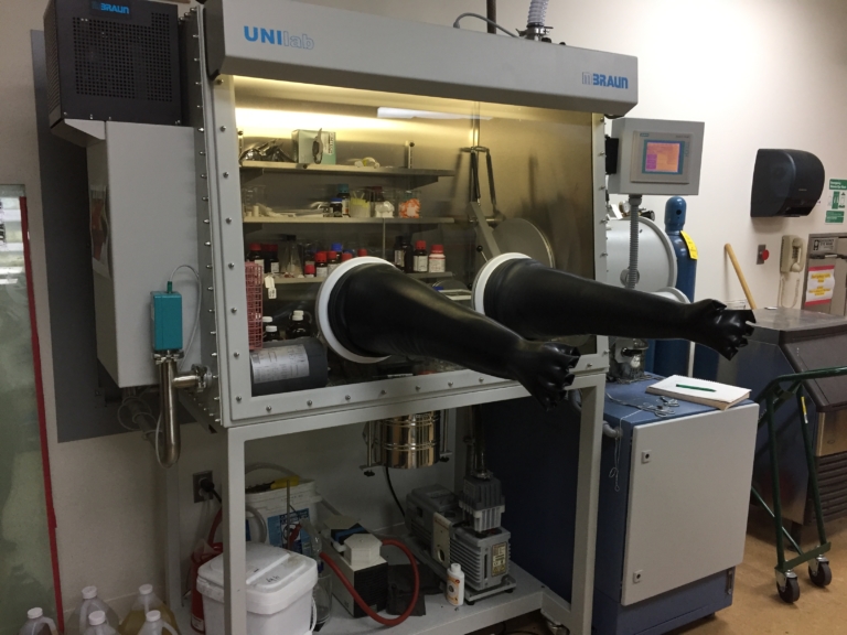 A laboratory glove box with black gloves extends from the front, containing equipment and chemicals inside on shelves.
