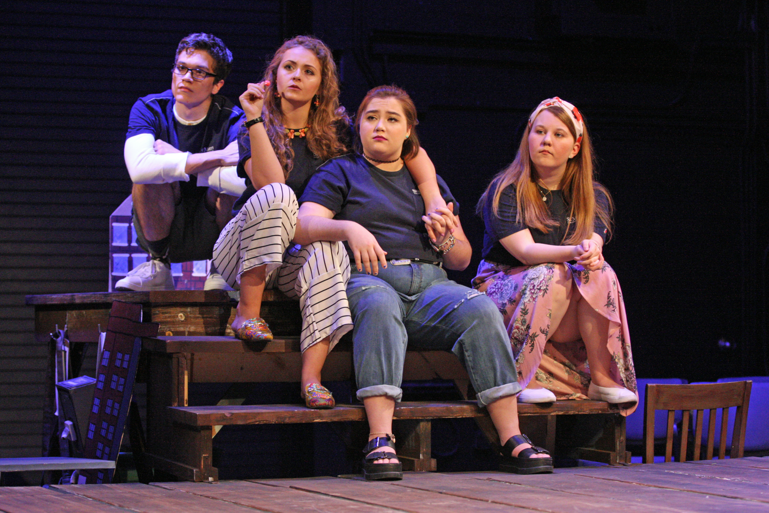 Four people sit closely together on a wooden stage set, wearing casual outfits, and look in various directions with thoughtful or serious expressions.