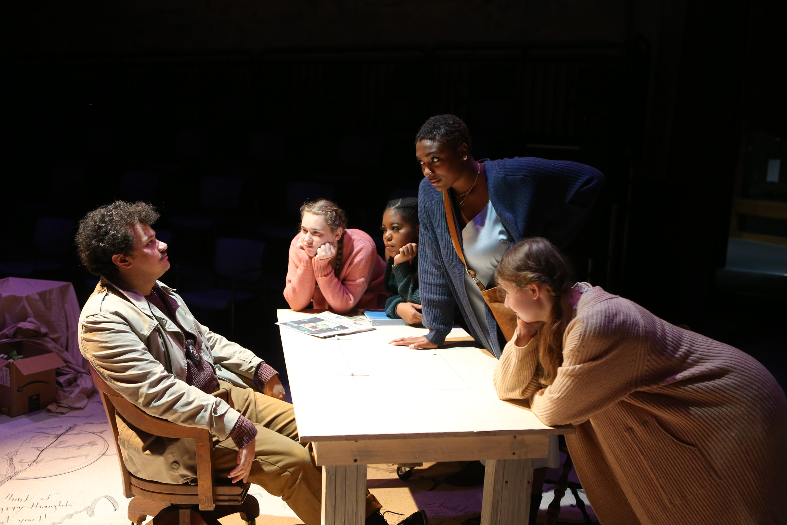 Five people are gathered around a table; one person sits while the others stand or lean in, all focused intently on the seated individual.