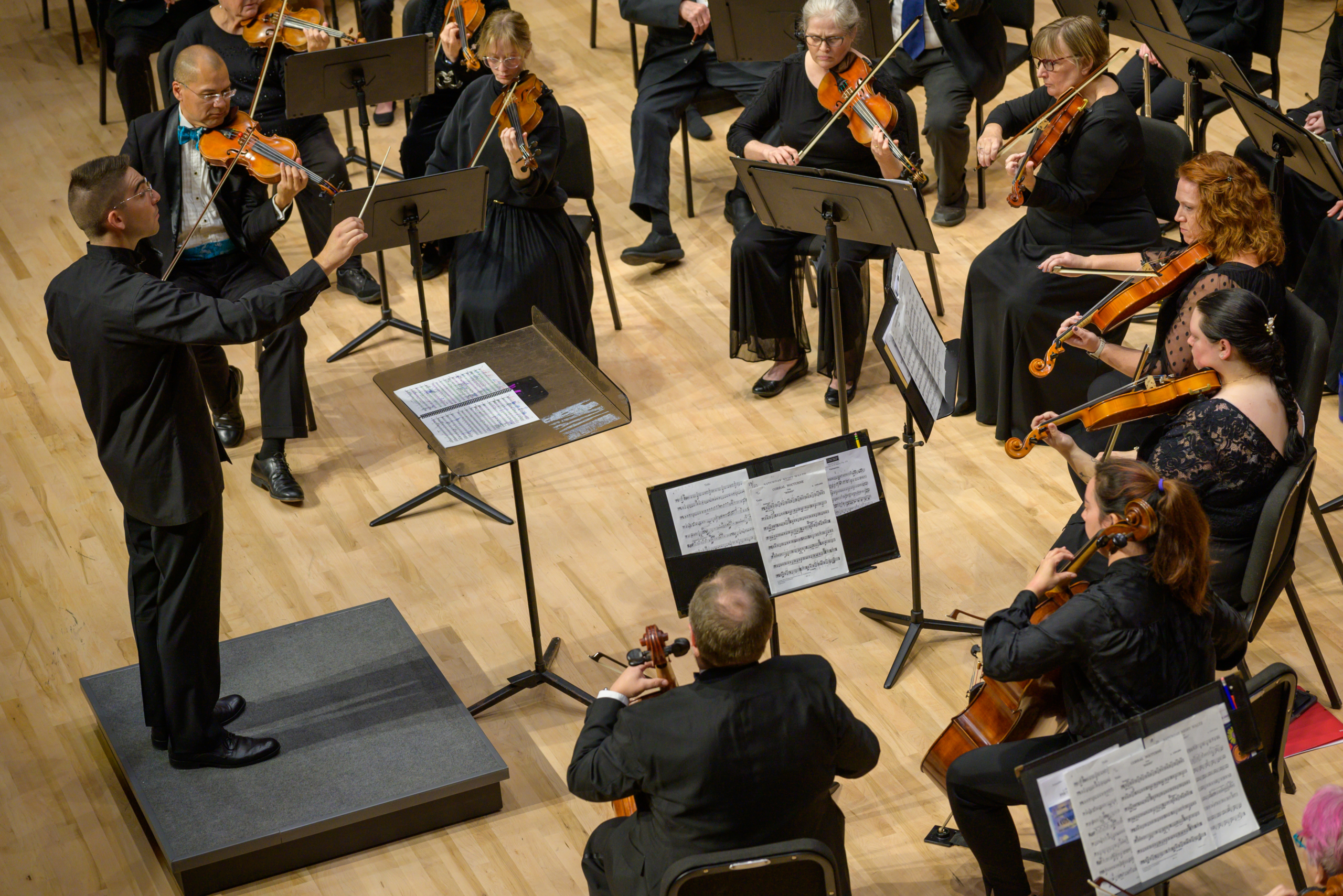A conductor leads an orchestra of violinists and violists, all dressed in black, performing on a wooden stage with sheet music on stands.