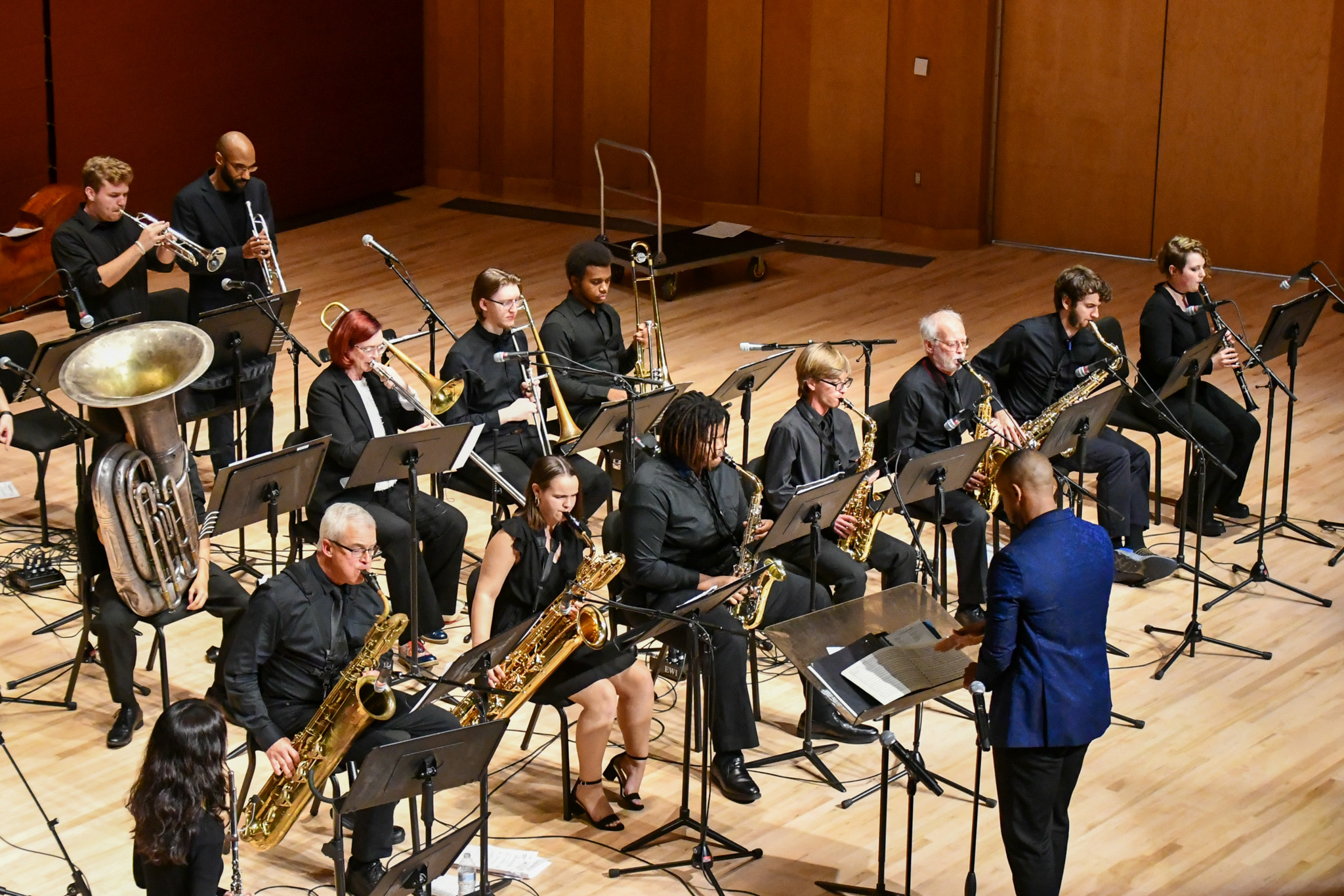 A jazz band performs on stage with musicians playing brass and woodwind instruments, led by a conductor in a blue jacket.