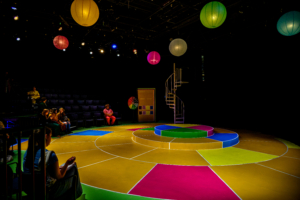 A round, multicolored stage with tiered platforms is set under hanging lanterns, with a small audience seated in a darkened theater.