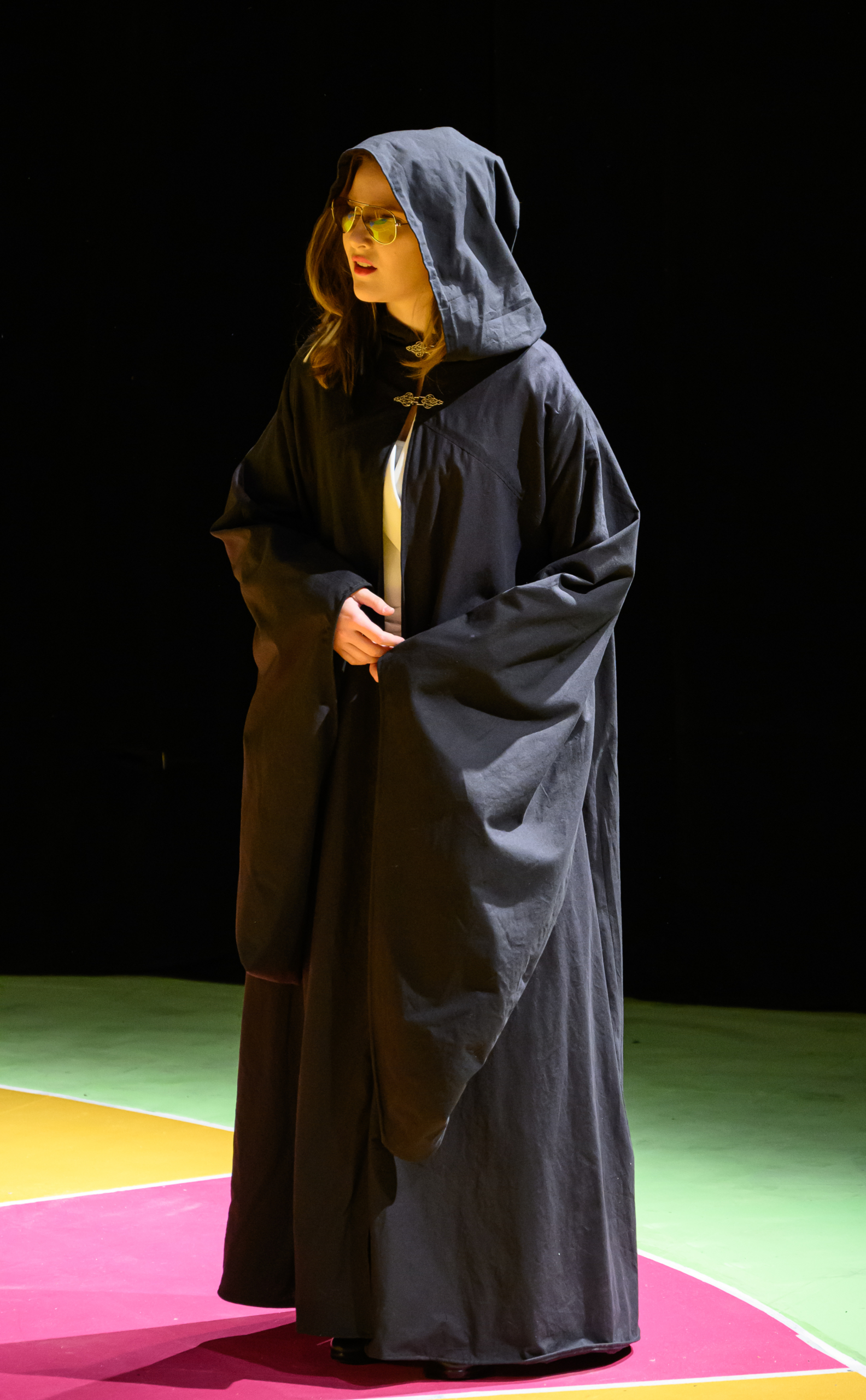 A person wearing a long black hooded robe stands on a multicolored stage with a dark background.