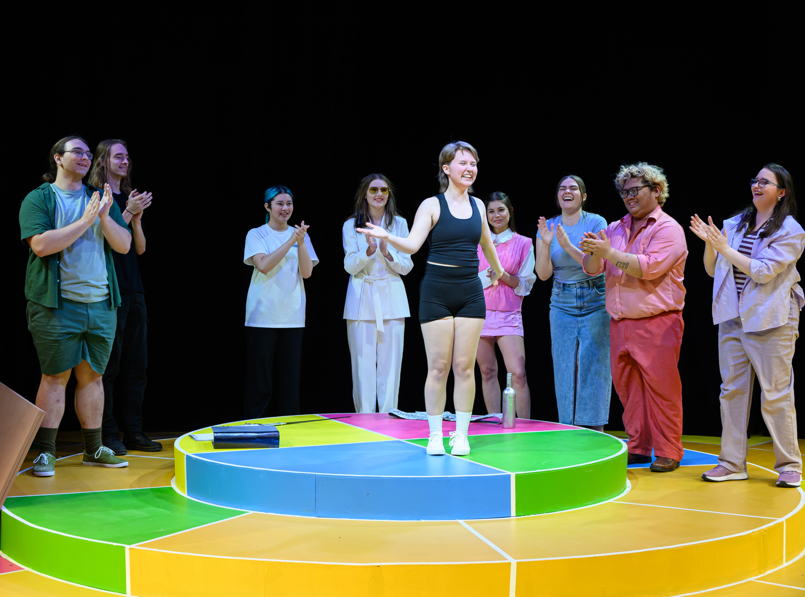A group of people stand on a colorful circular stage, clapping and smiling as one person in athletic wear stands at the center.