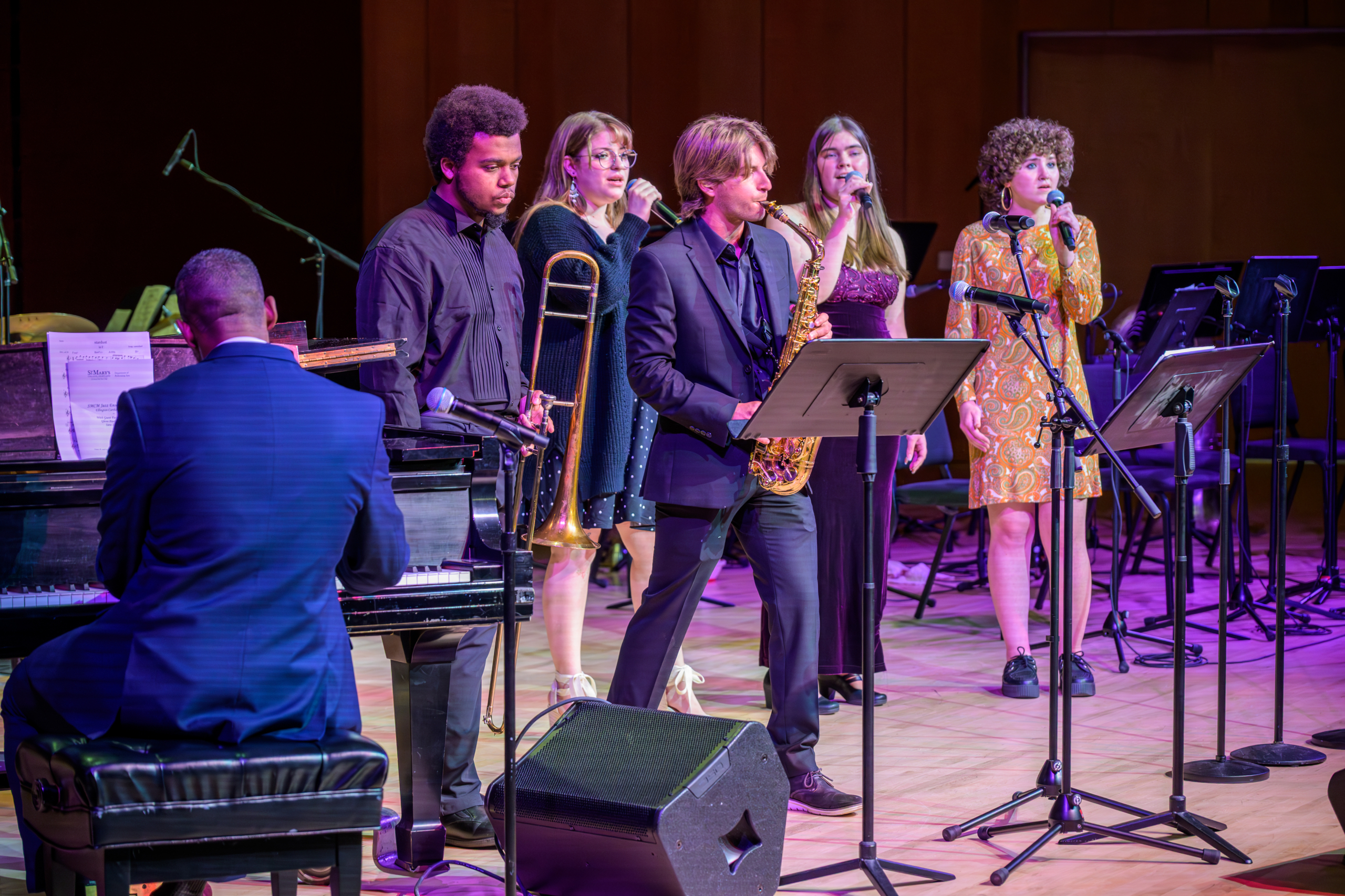 A jazz band performs on stage; a pianist, trombonist, saxophonist, and three vocalists stand under stage lights with music stands and microphones.