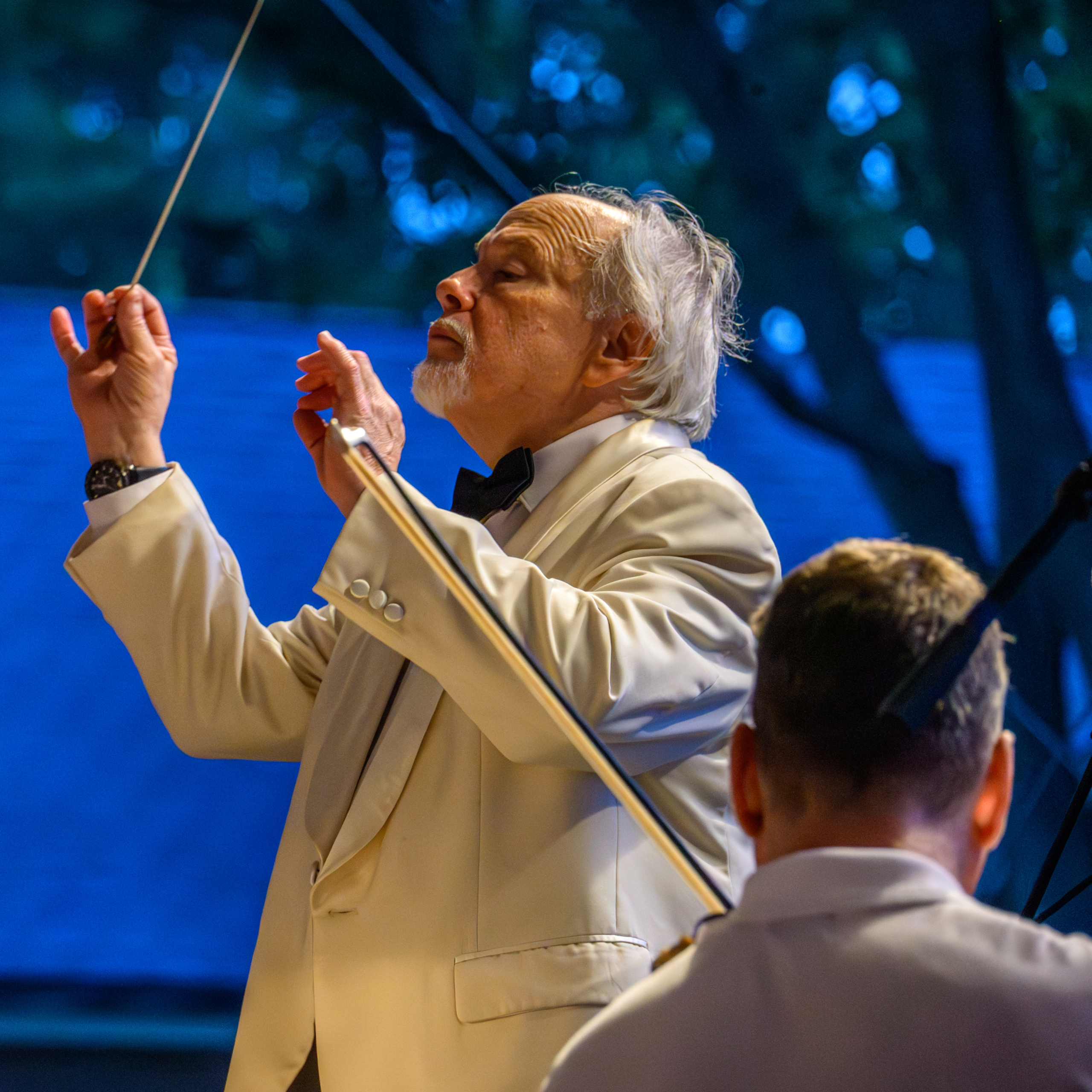 An older man in a white suit conducts an orchestra outdoors, raising a baton with both hands as another musician sits in the foreground.