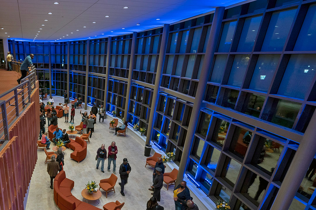 People gather and converse in a spacious, modern lobby with large glass windows and blue accent lighting in the evening.