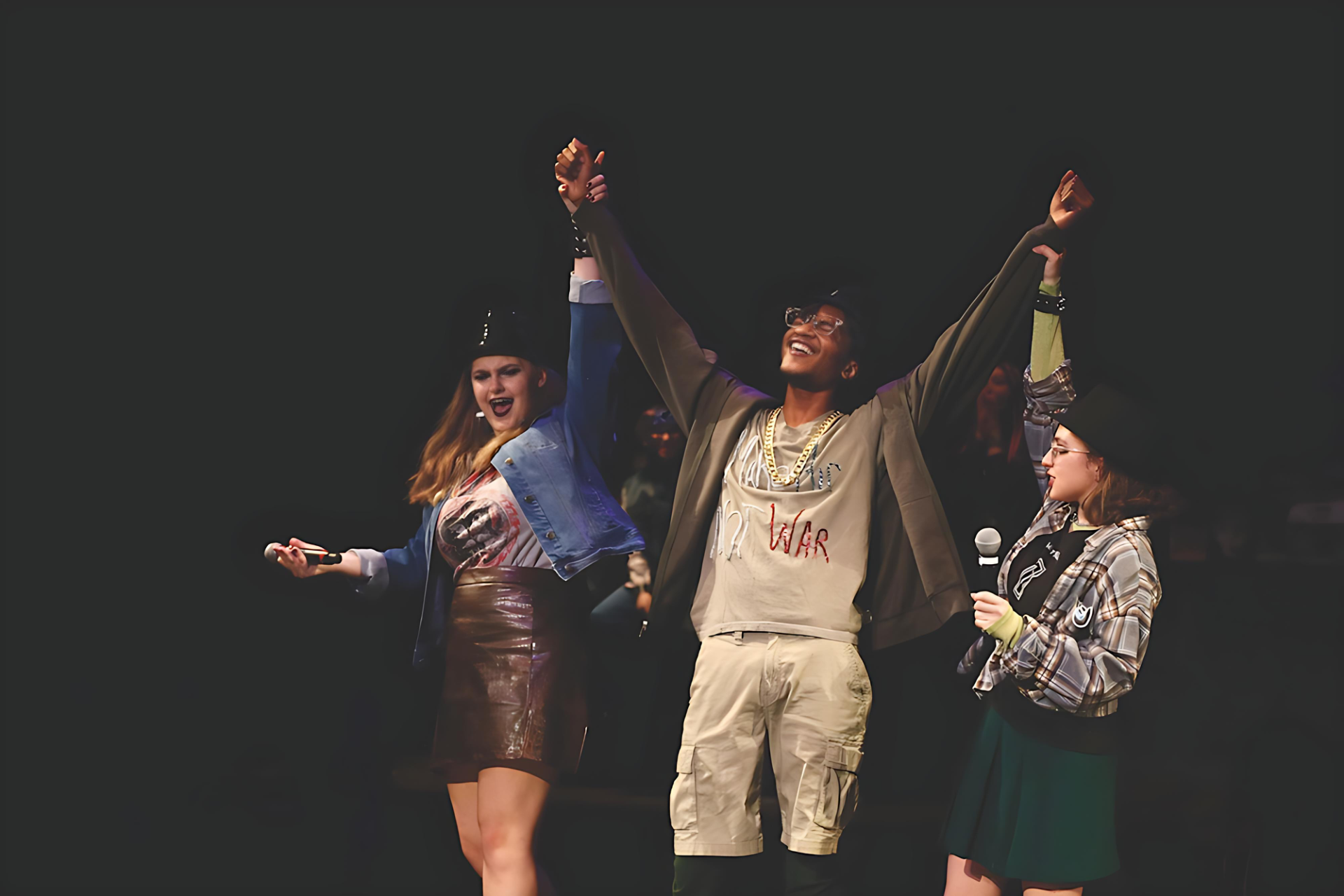 Three people stand on stage with hands raised in celebration; one holds a microphone and another wears a shirt with "NO WAR" written on it.