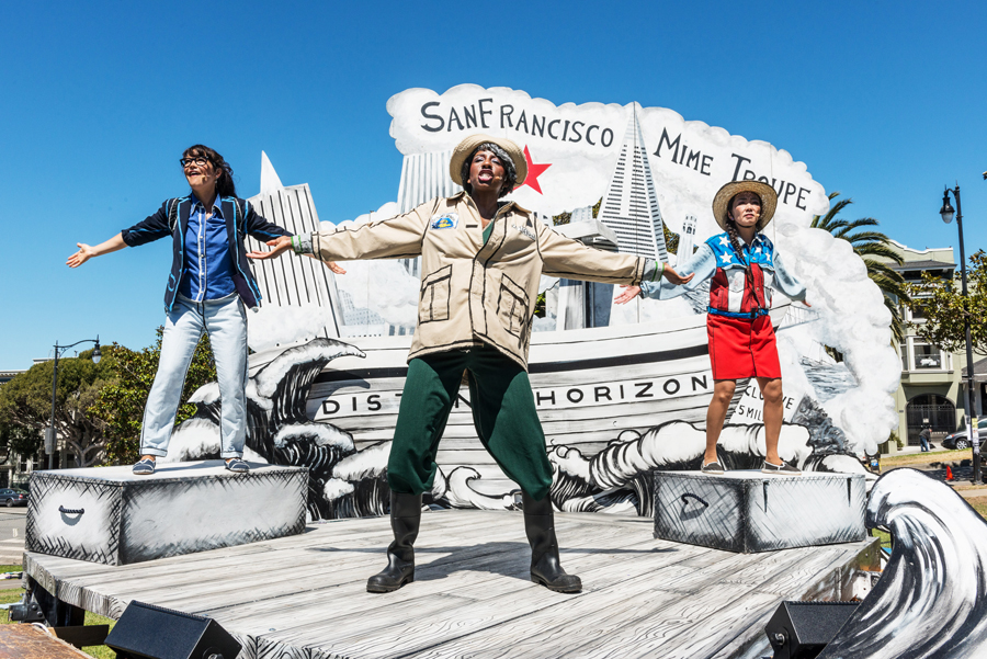 Three performers stand with arms outstretched on a stage with a painted San Francisco backdrop, under a sign that reads "San Francisco Mime Troupe.