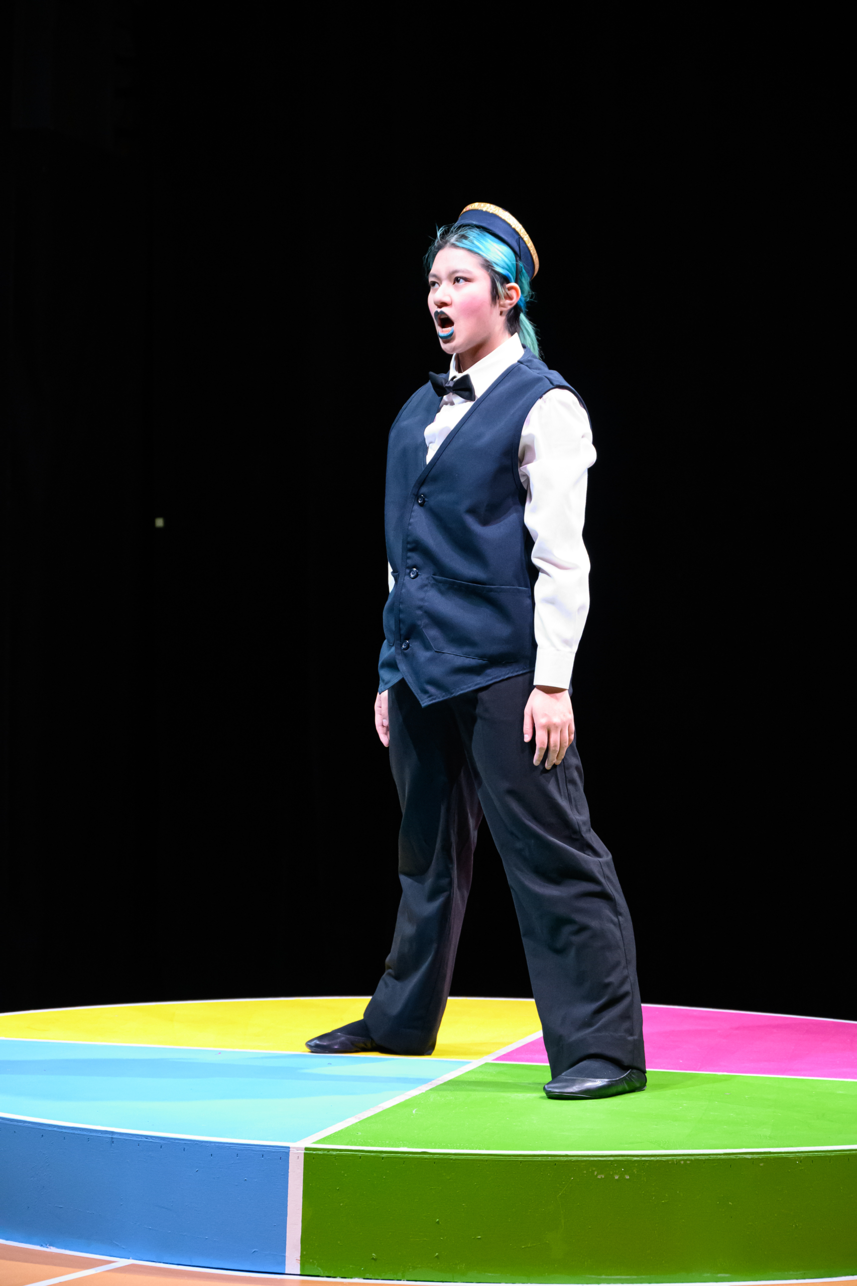 A person in a vest and bowtie stands on a colorful circular platform, singing or speaking with an expressive face against a dark background.