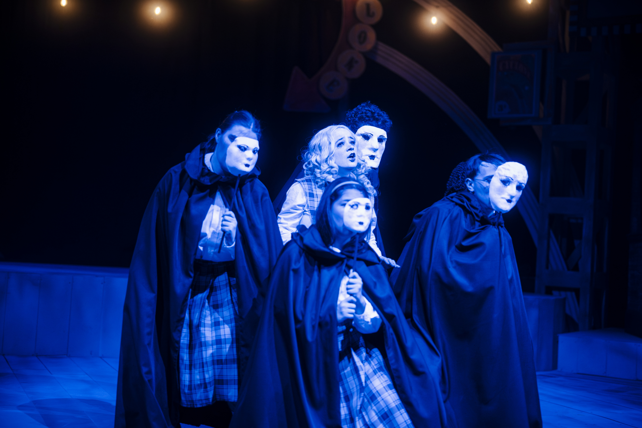 Five performers wearing white masks and dark capes stand closely together on stage under blue lighting, with theatrical set pieces and string lights visible in the background.