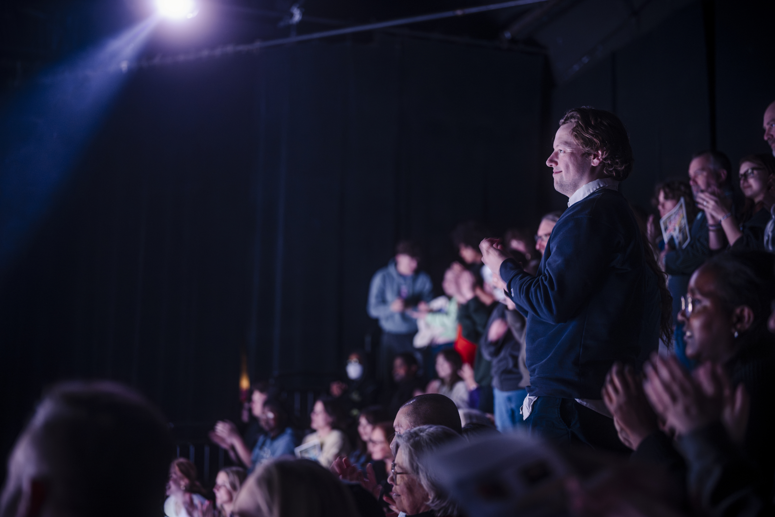 Audience members stand and applaud in a dimly lit theater, with a spotlight illuminating part of the crowd.
