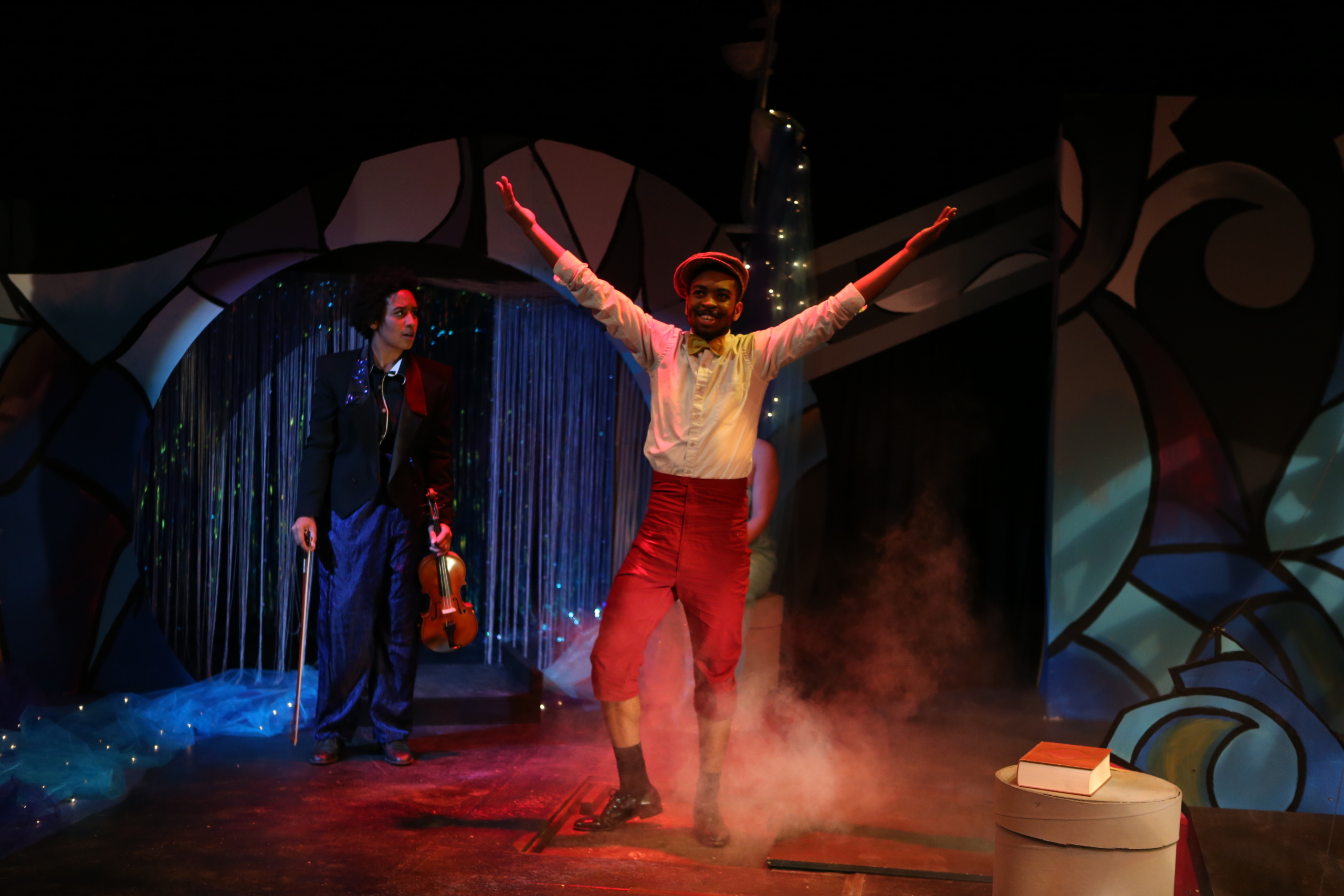 A performer in a white shirt and red pants stands with arms raised center stage, while another person with a violin stands behind him under theatrical lighting and stage decorations.