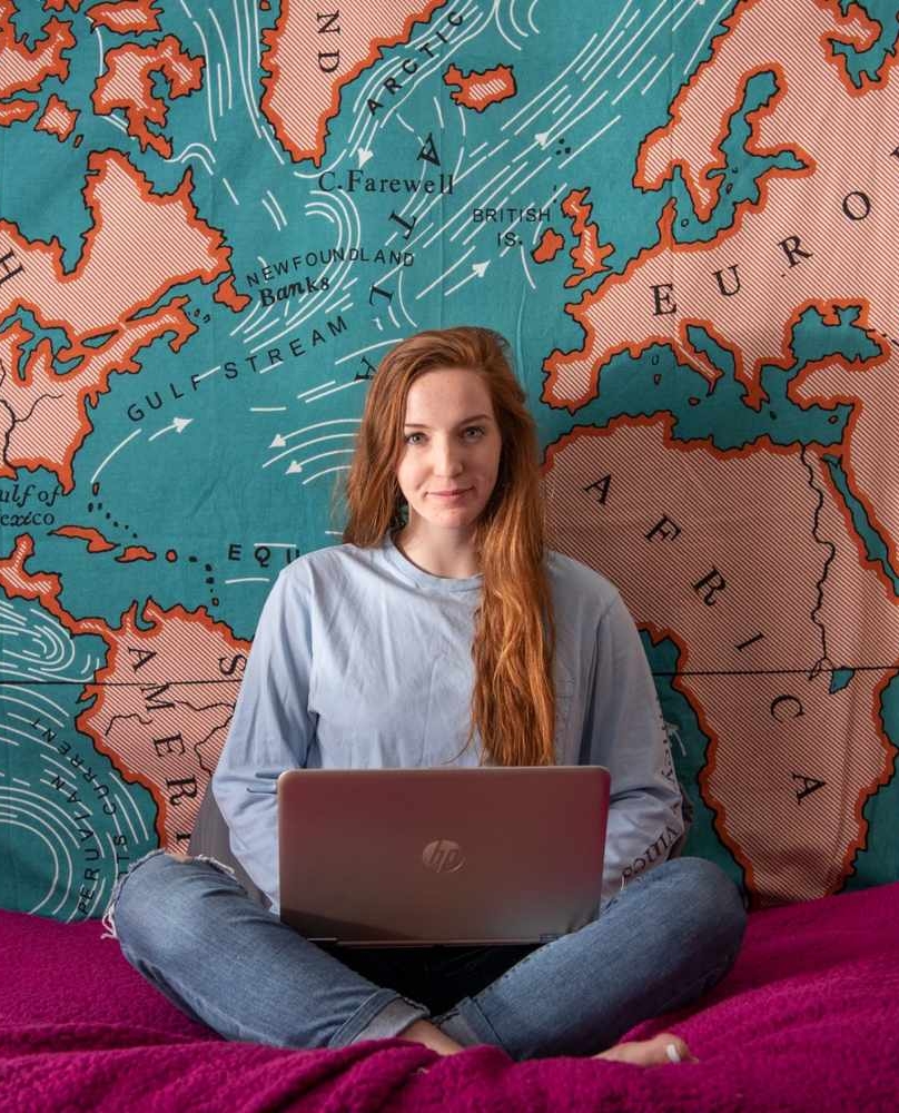 A person with long red hair sits cross-legged on a bed using a laptop, with a large world map tapestry hanging on the wall behind them.