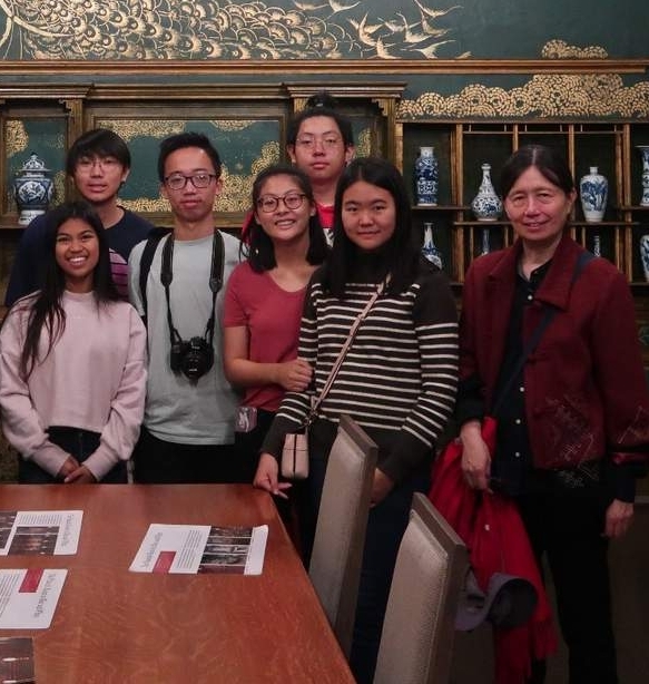A group of ten people pose for a photo in a decorative room with a gold mural of birds on a green wall and shelves with blue and white vases behind them.