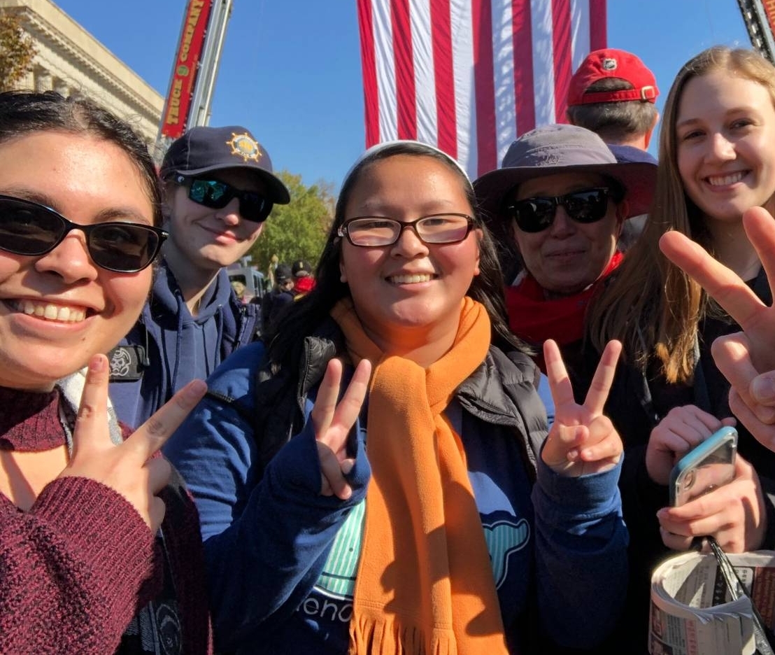 A group of people smiles and holds up peace signs in front of a large hanging American flag at an outdoor event.