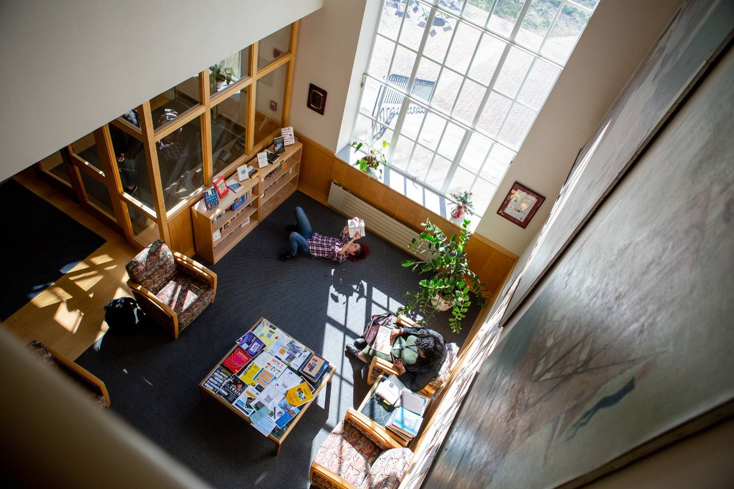 A person lies on the floor reading a book in a sunlit library lounge with armchairs, tables, bookshelves, and large windows.