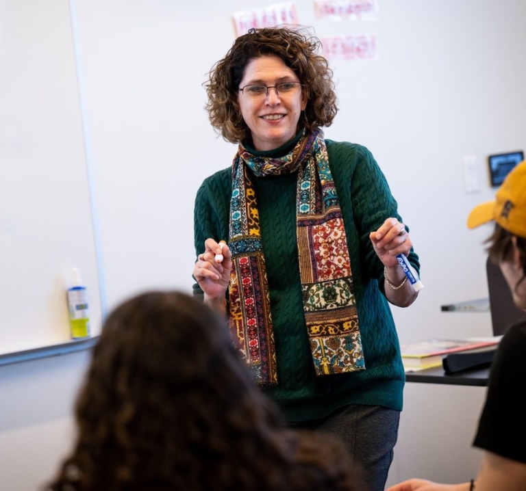 A woman wearing glasses and a patterned scarf stands and speaks in front of a whiteboard to students in a classroom.