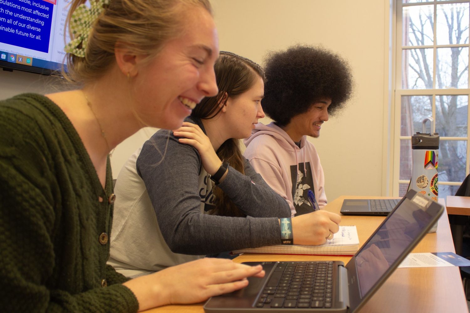 Three students sit at desks using laptops and taking notes in a classroom with a window and a wall-mounted screen in the background.