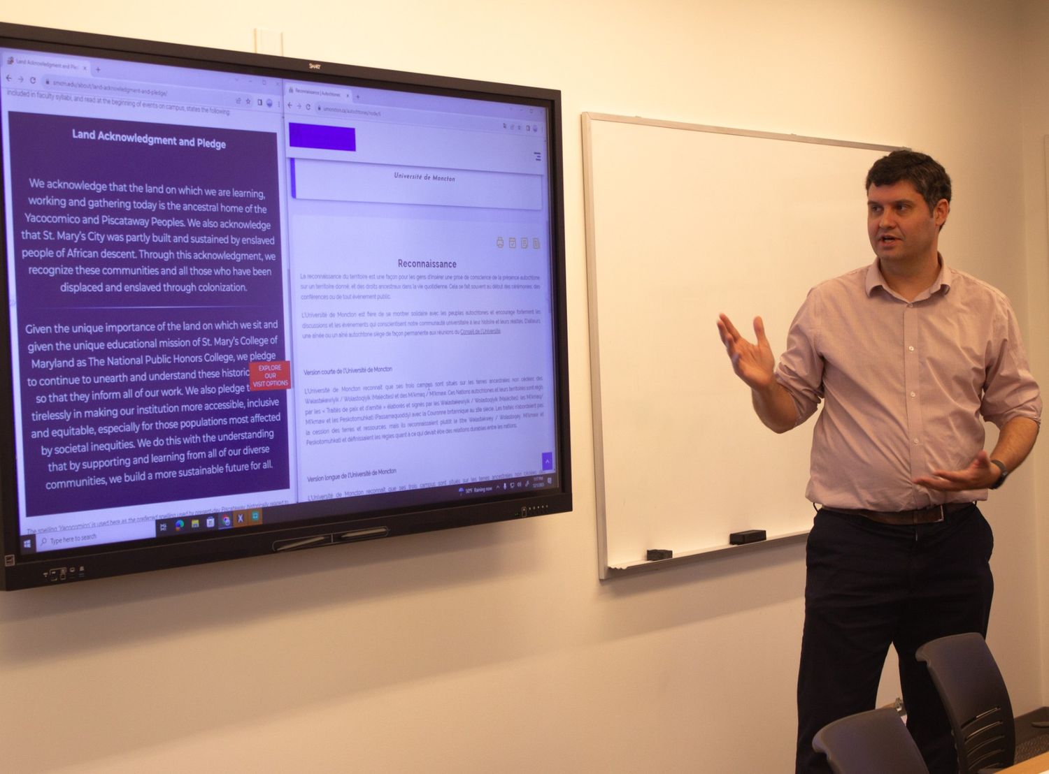 A man stands next to a large screen displaying a land acknowledgement and text, gesturing with one hand while presenting in a classroom.